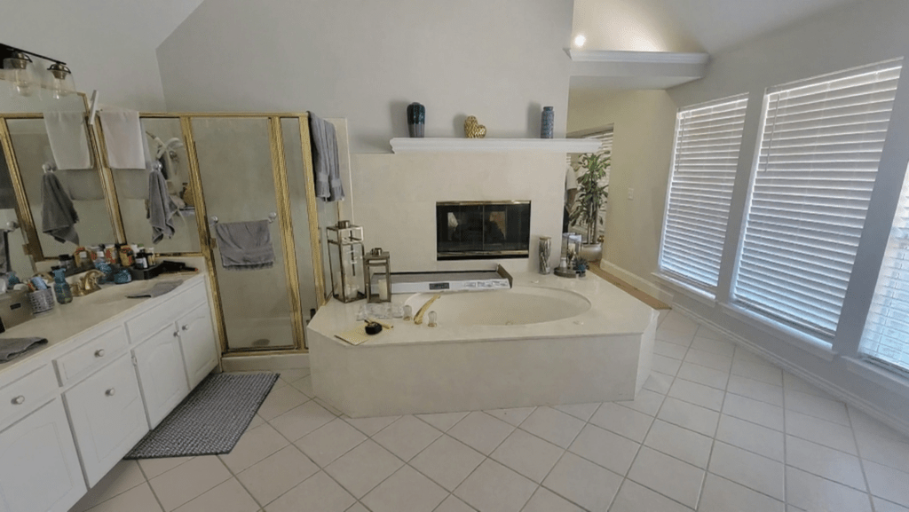 Before master bath remodel Colleyville TX showing oversized garden tub with gold faucet, dated fireplace surround, gold frame shower enclosure and basic white tile — transformed by I Love My Bathroom