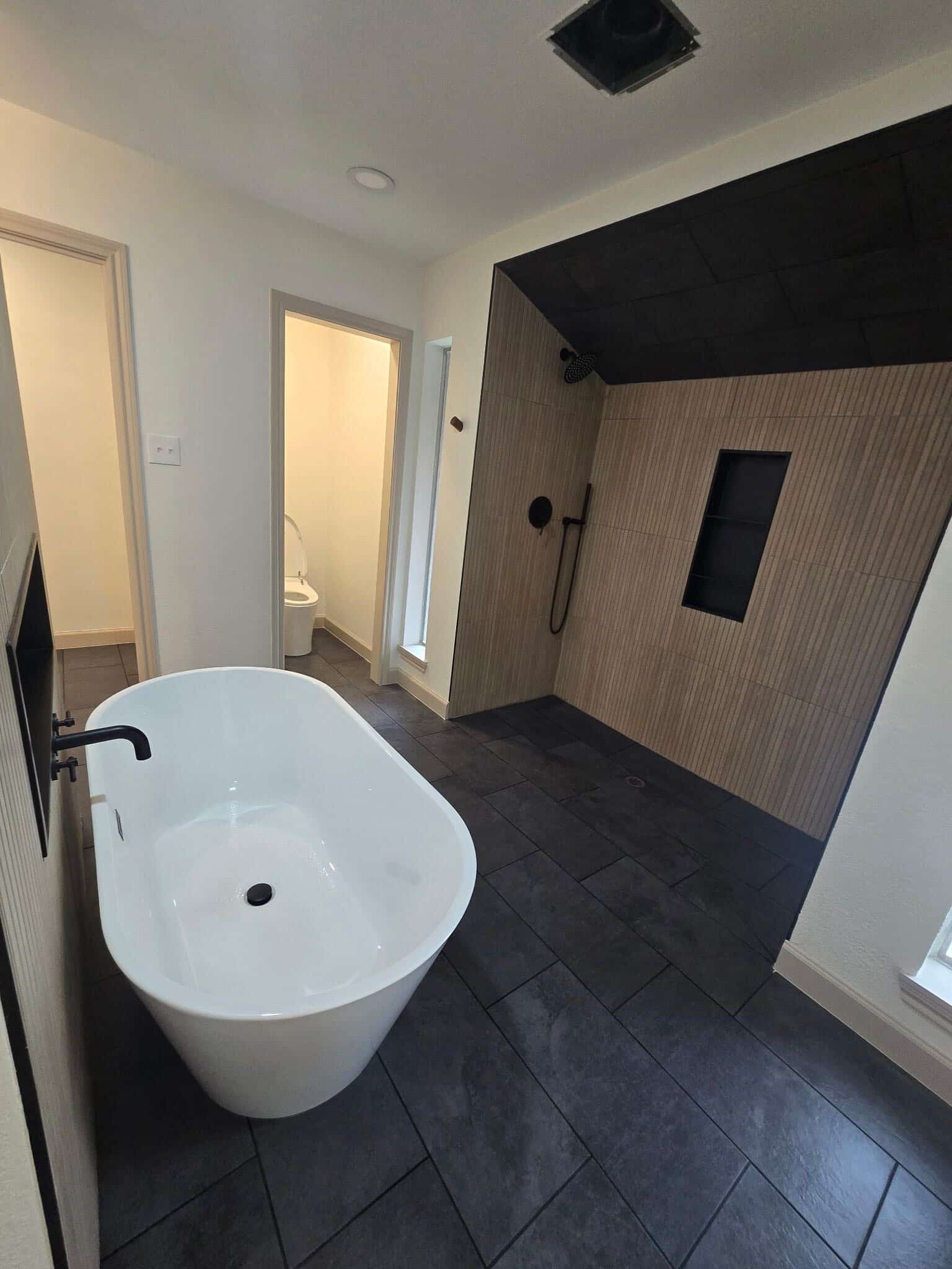 Freestanding soaking tub DFW bathroom remodel featuring open wet room walk-in shower with warm wood look vertical tile matte black ceiling dark charcoal floor tile and matte black hardware by I Love My Bathroom