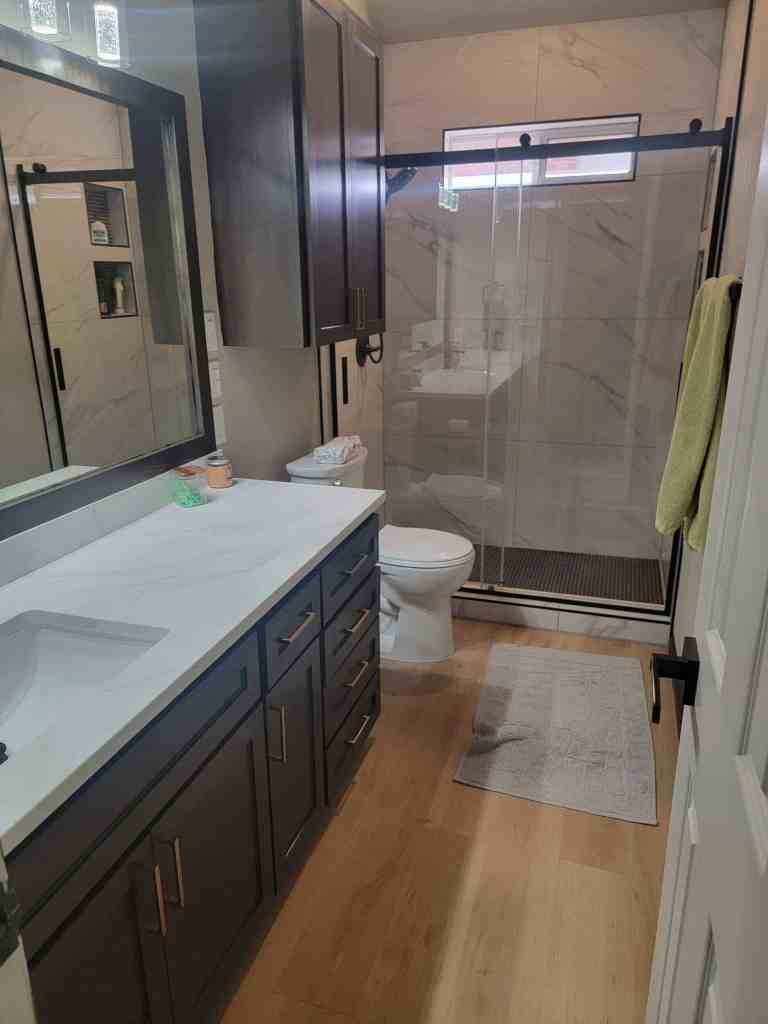 Full guest bathroom remodel Denton TX featuring dark navy vanity with quartz countertop, marble porcelain shower tile, frameless glass enclosure with matte black hardware, and luxury vinyl plank flooring by I Love My Bathroom