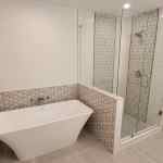 Master bath remodel Grapevine TX featuring freestanding soaking tub with wall mount filler geometric pattern tile accent wall walk-in shower with frameless glass and wood look floor tile by I Love My Bathroom